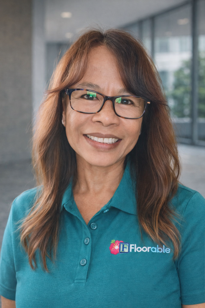 Woman smiling in a teal Floorable Hawaii polo shirt, showcasing expertise in flooring solutions, with a blurred office background.