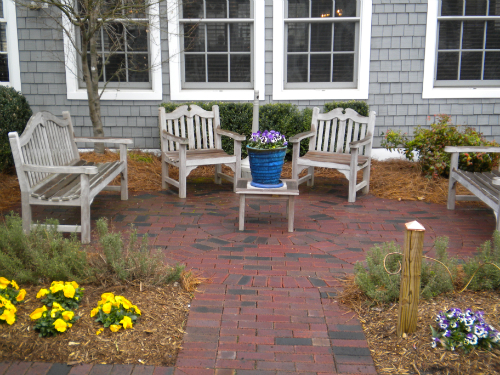 Brick-paved seating area with wooden benches, central flower pot, and landscaped greenery