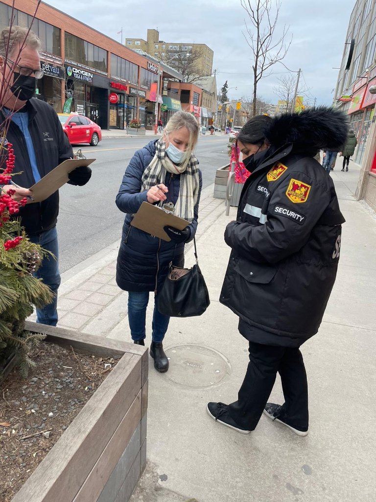 Security guard assisting two individuals with clipboard on a city street, emphasizing professional security services and community engagement.