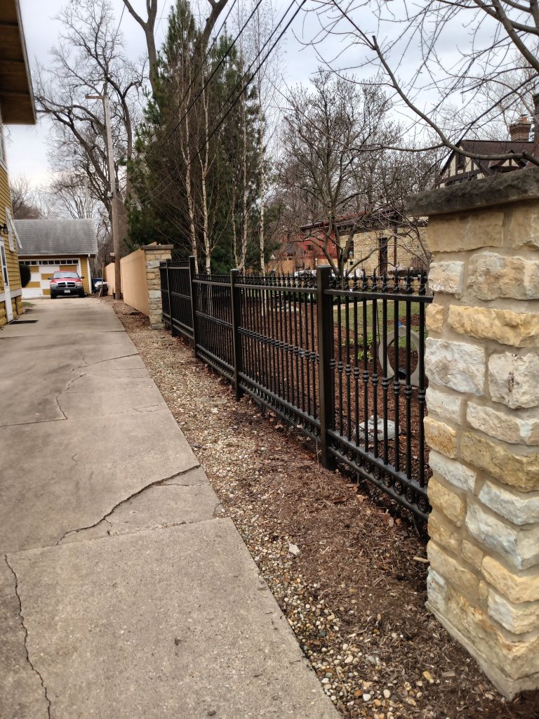 Extended black ornamental iron perimeter fence with decorative pickets and collars spanning full residential property line between stone masonry columns