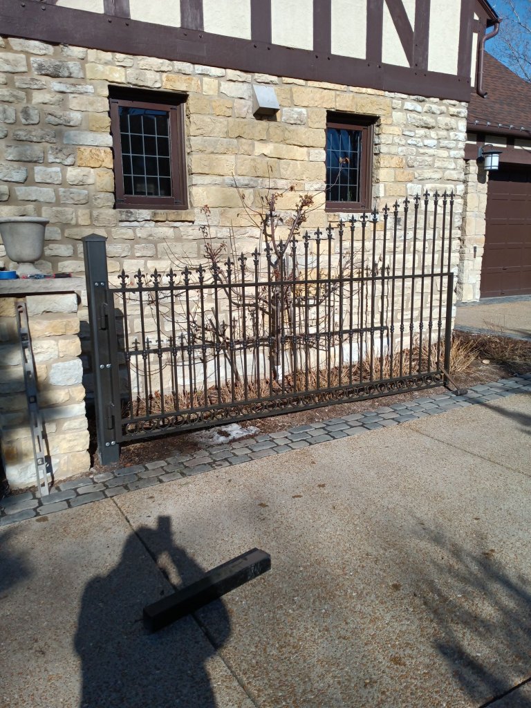Custom ornamental iron driveway gate panel with fleur-de-lis finials and decorative scrollwork being installed at residential estate with stone masonry pillars
