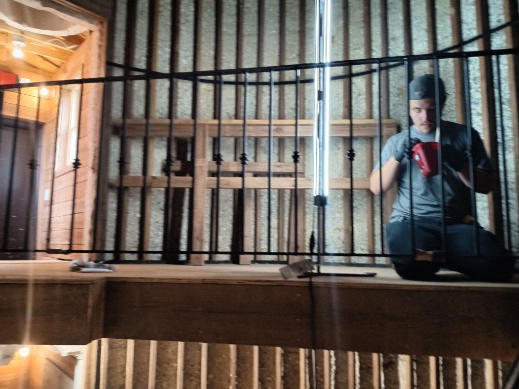 On-site welder installing curved iron stair railing with decorative balusters on wood staircase inside converted grain silo tower, Miller welder visible