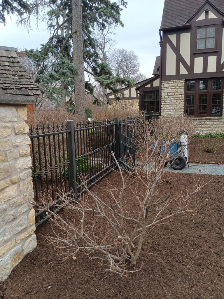Custom black ornamental iron fence panel with integrated walk gate installed at corner of residential property with stone columns and Tudor-style home