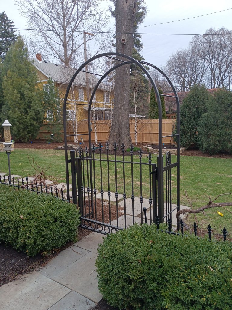 Custom fabricated ornamental iron garden entry gate with decorative arched overhead structure, spear-top finials, and fleur-de-lis accents installed at residential property