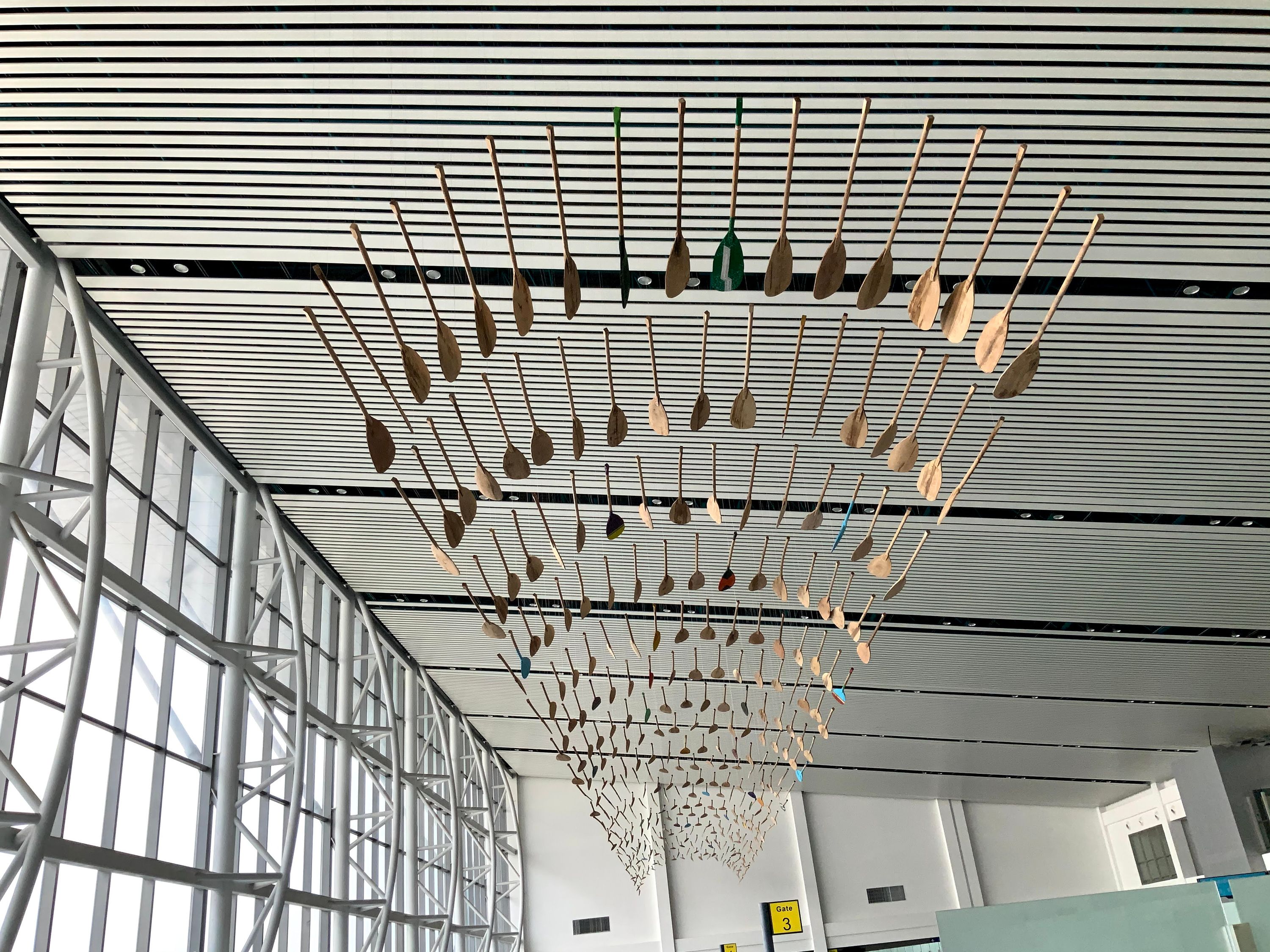 Hundreds of suspended wooden oars hung in a wave formation beneath a slatted ceiling in a glass-walled hall