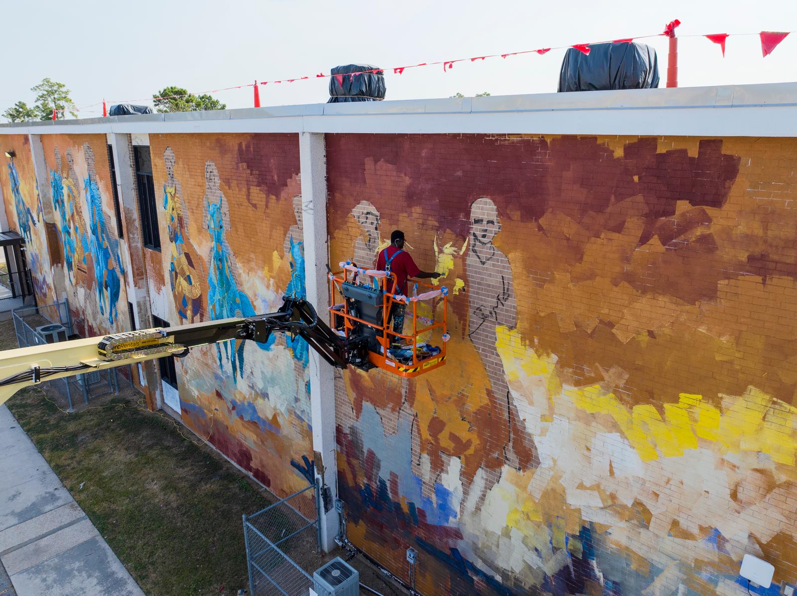 Aerial view of Adeodunfa on a boom lift painting the Free Spirits, Golden Hearts mural on a brick building