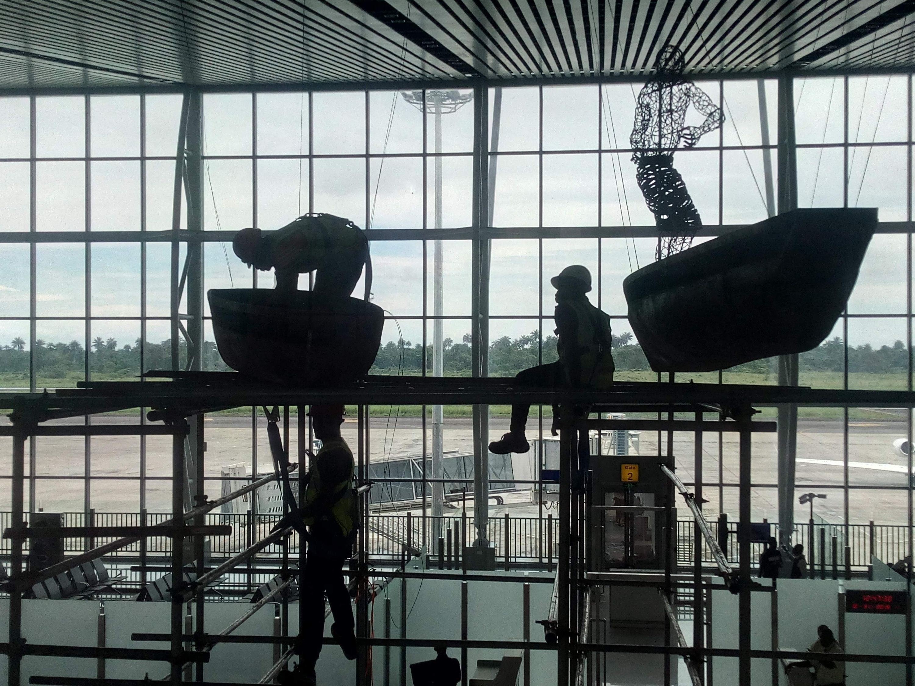 Installation crew on scaffolding hanging suspended boat and figure sculptures in front of a tall glass wall