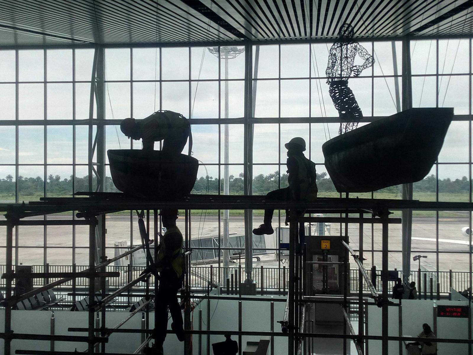 Installation crew on scaffolding hanging suspended boat and figure sculptures in front of a tall glass wall
