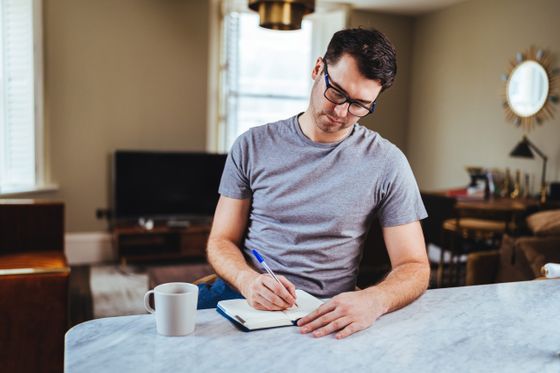 Dean writing in a notebook at a table at home, a mug beside him