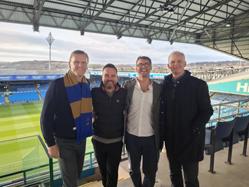 Four people in a football stand, the pitch and the far stand behind them
