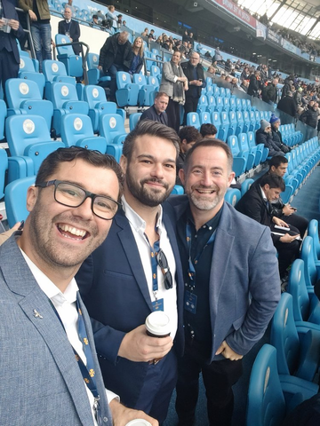 Three people in the stands at the Etihad, the blue seats filling up behind them