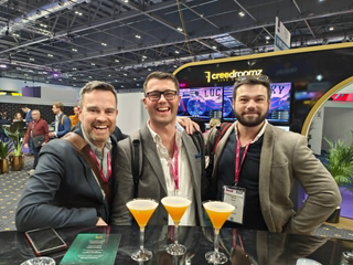 Three people in suits with drinks in hand at a trade-show stand, the hall lit behind them