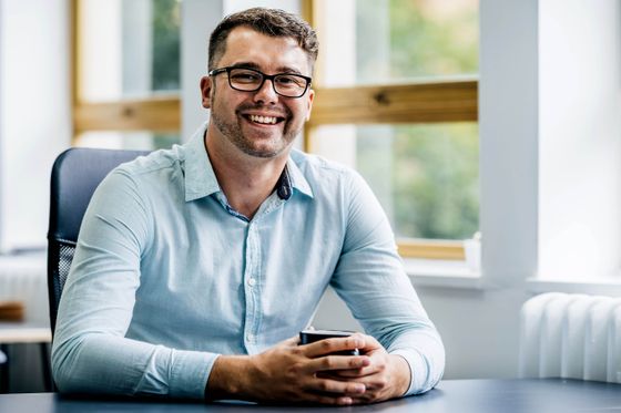 A Trusted Media photograph: someone at a desk by a window with a coffee, looking up from the work