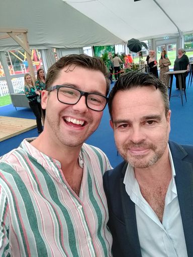 Two people taking a selfie, both grinning, an event behind them