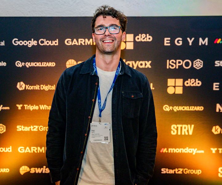 Dean Lynn, in a dark jacket and a conference lanyard, smiling at the camera in front of an event backdrop tiled with sponsor logos