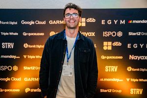 Dean in a dark jacket and lanyard in front of an event backdrop tiled with sponsor logos