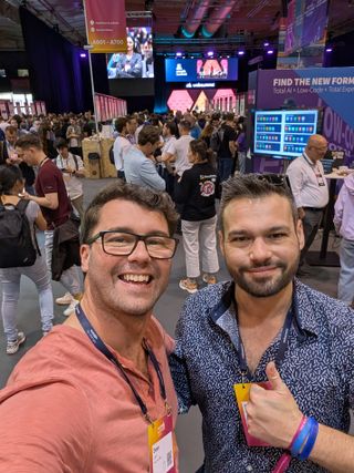 A selfie of two people on a busy exhibition floor, a huge pink stage screen and crowds behind