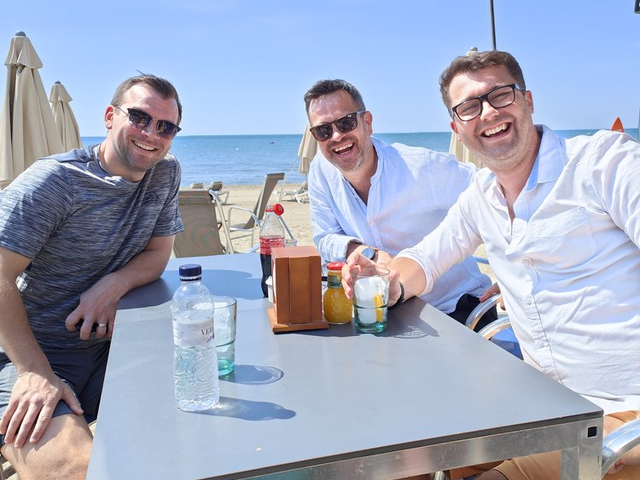Dean and two friends laughing around a table at a beach bar in Spain, the sea and a clear blue sky behind them.