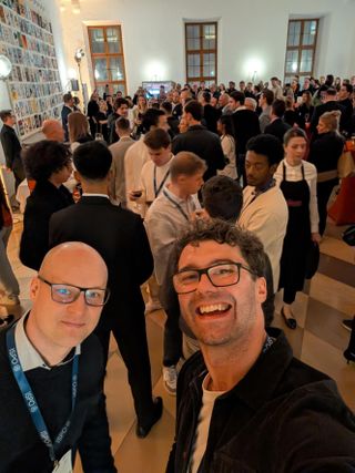 A selfie of two people at a busy evening drinks reception in a tall white-walled hall