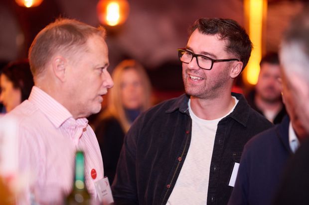 Two people in conversation at a crowded, warmly lit bar, others talking behind them