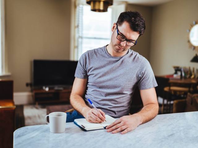 Dean writing in a notebook at a marble kitchen island in a bright Stamford apartment, a mug of coffee beside him.